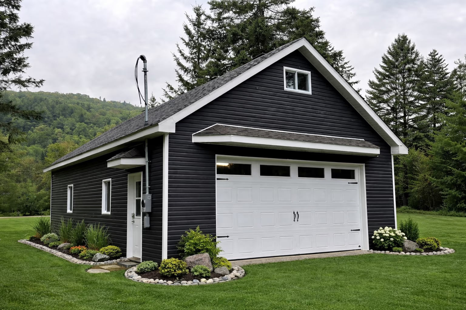 Black and white detached garage with large white garage door, dark exterior siding, shingle roof, green lawn and flowered landscaping