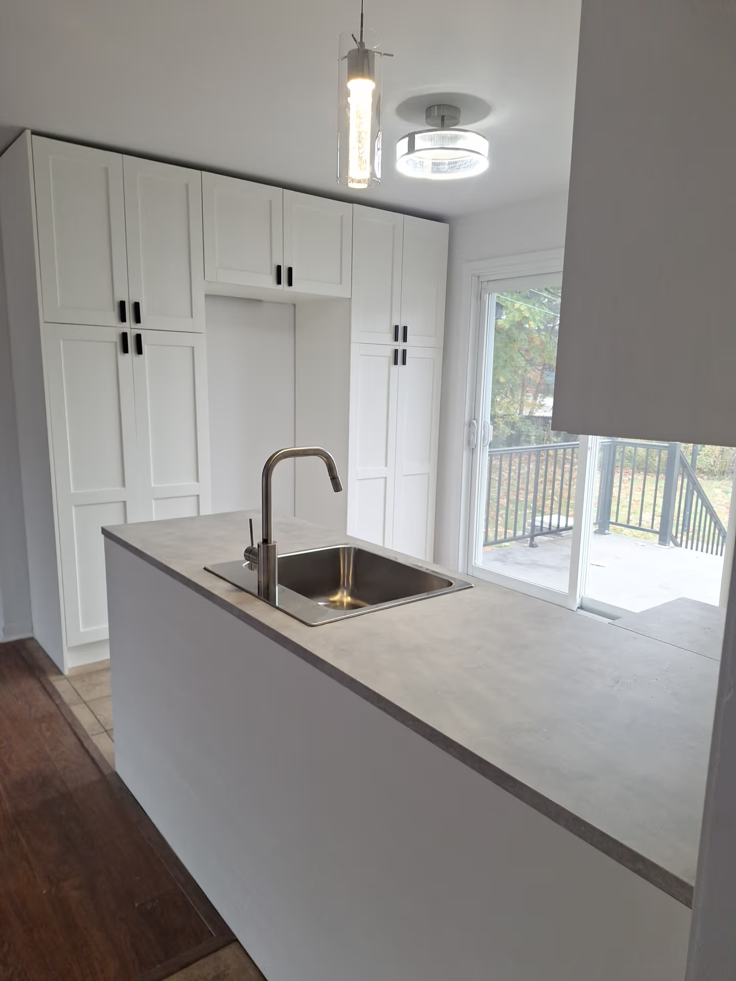 Cuisine lumineuse avec armoires blanches, grand comptoir gris, évier intégré, robinet chromé, porte patio vitrée et luminaires modernes