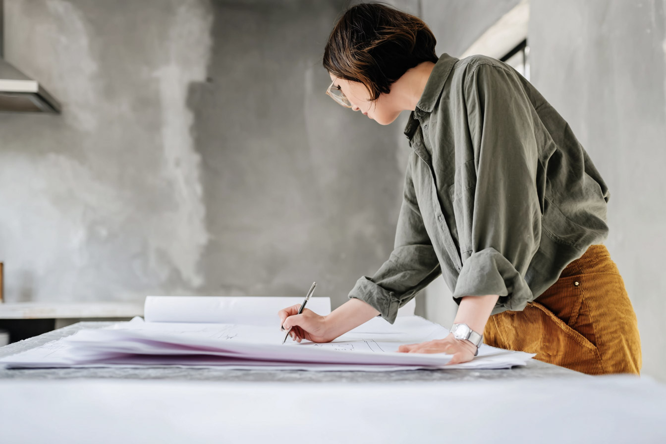 Architect or interior designer drawing on renovation plans in an unfinished space, with raw concrete wall and large worktable.