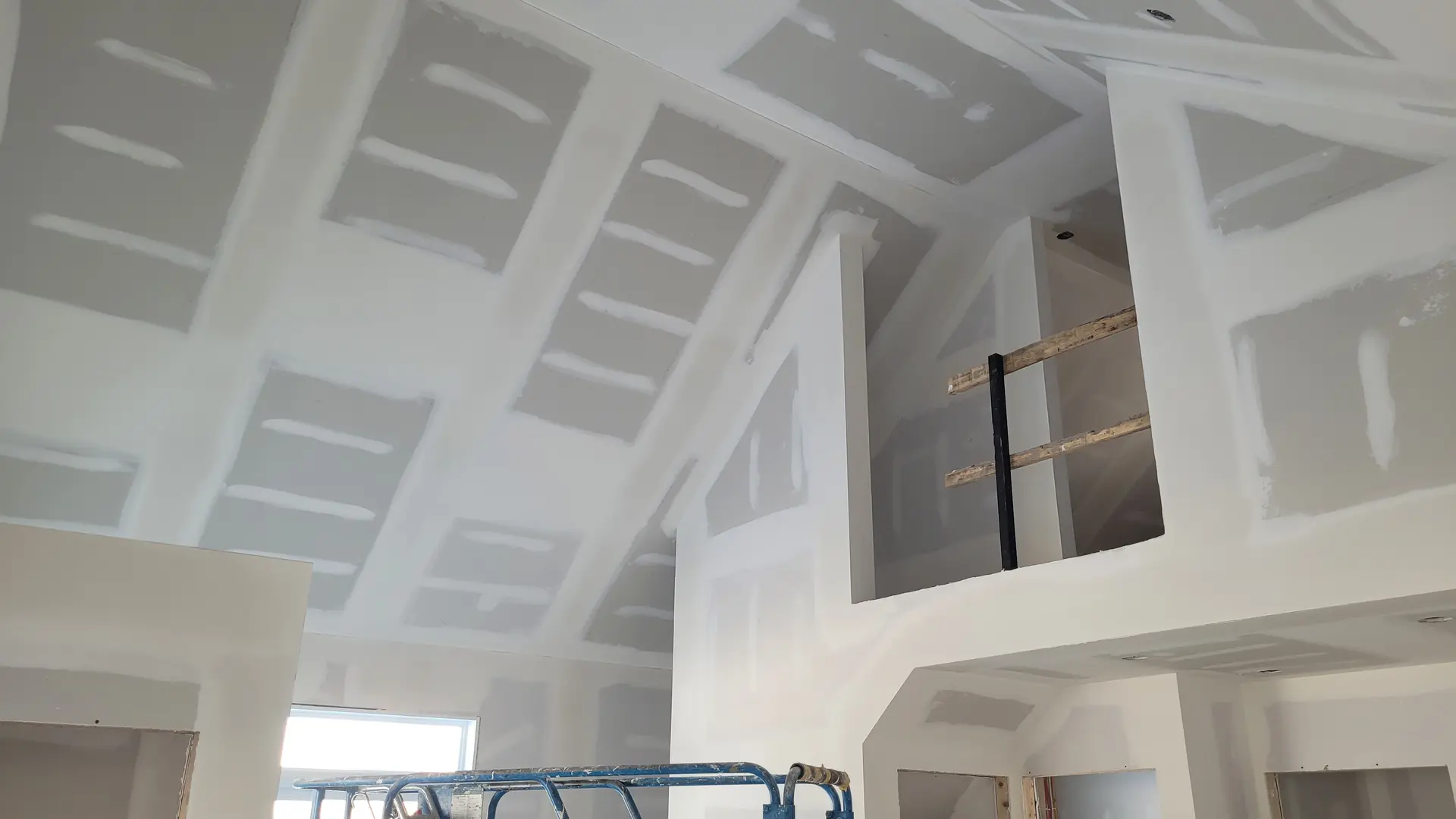 Drywall installation and finishing work on walls and cathedral ceilings of a house under construction.