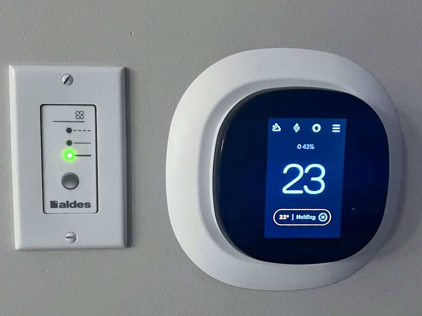 Thermostat numérique affichant une température de 23 °C à côté d’un panneau de contrôle de ventilation Aldes, fixé sur un mur gris.
