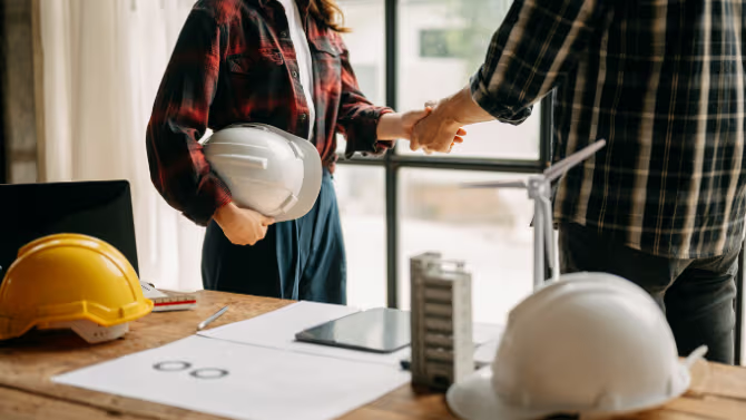 Deux professionnels de la construction se serrant la main près de plans, casques de sécurité, ordinateur portable et maquette de bâtiment.