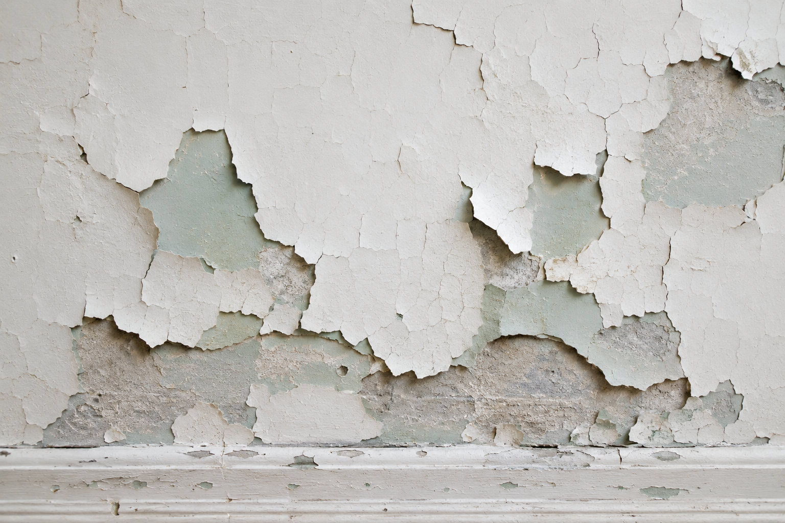 Old interior wall with chipped white paint revealing pale green layers and a deteriorated plaster substrate.