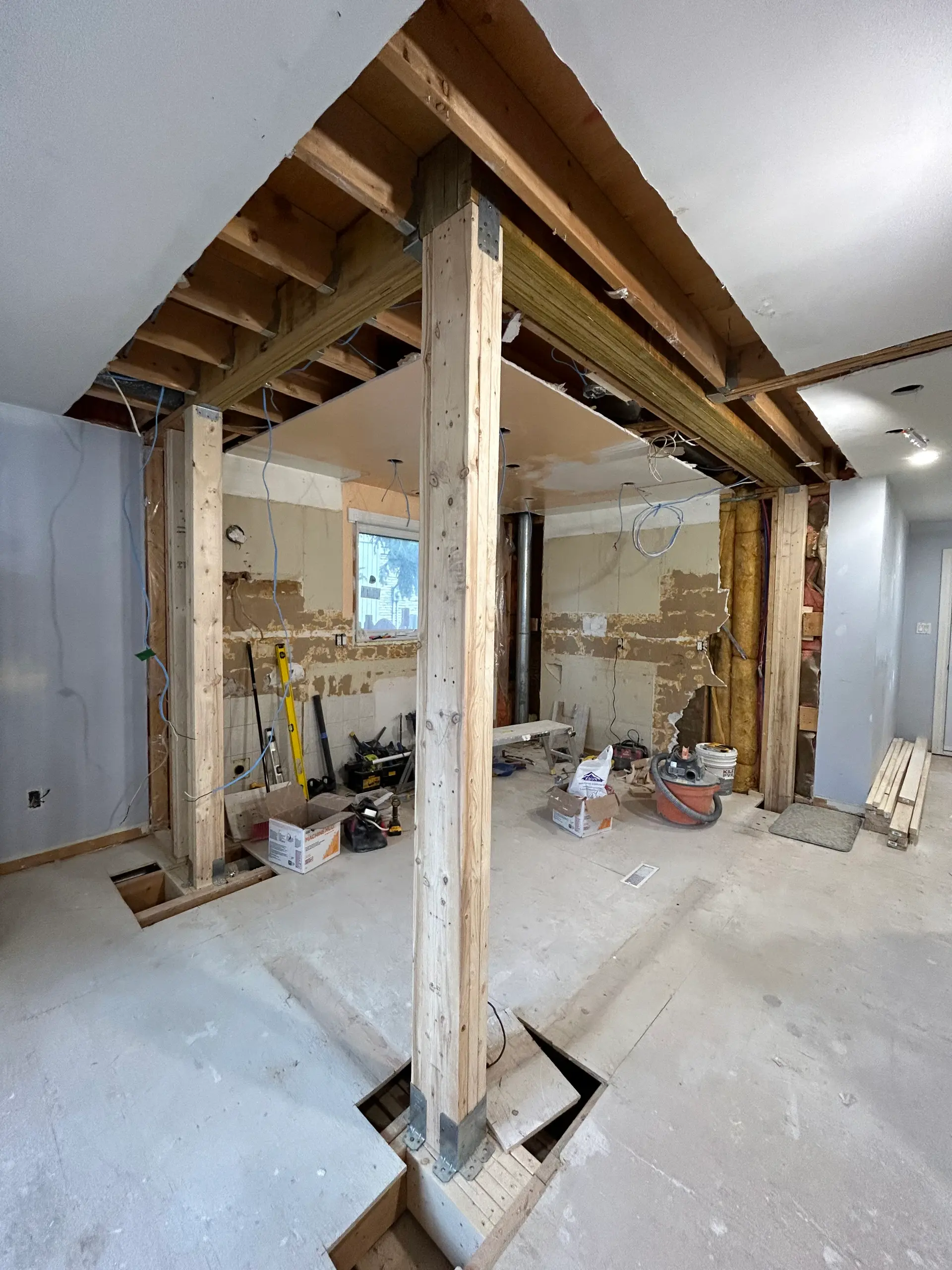 Intérieur de maison en rénovation majeure avec plafond ouvert, poutres et poteaux structuraux en bois, câblage apparent et murs partiellement démolis. 