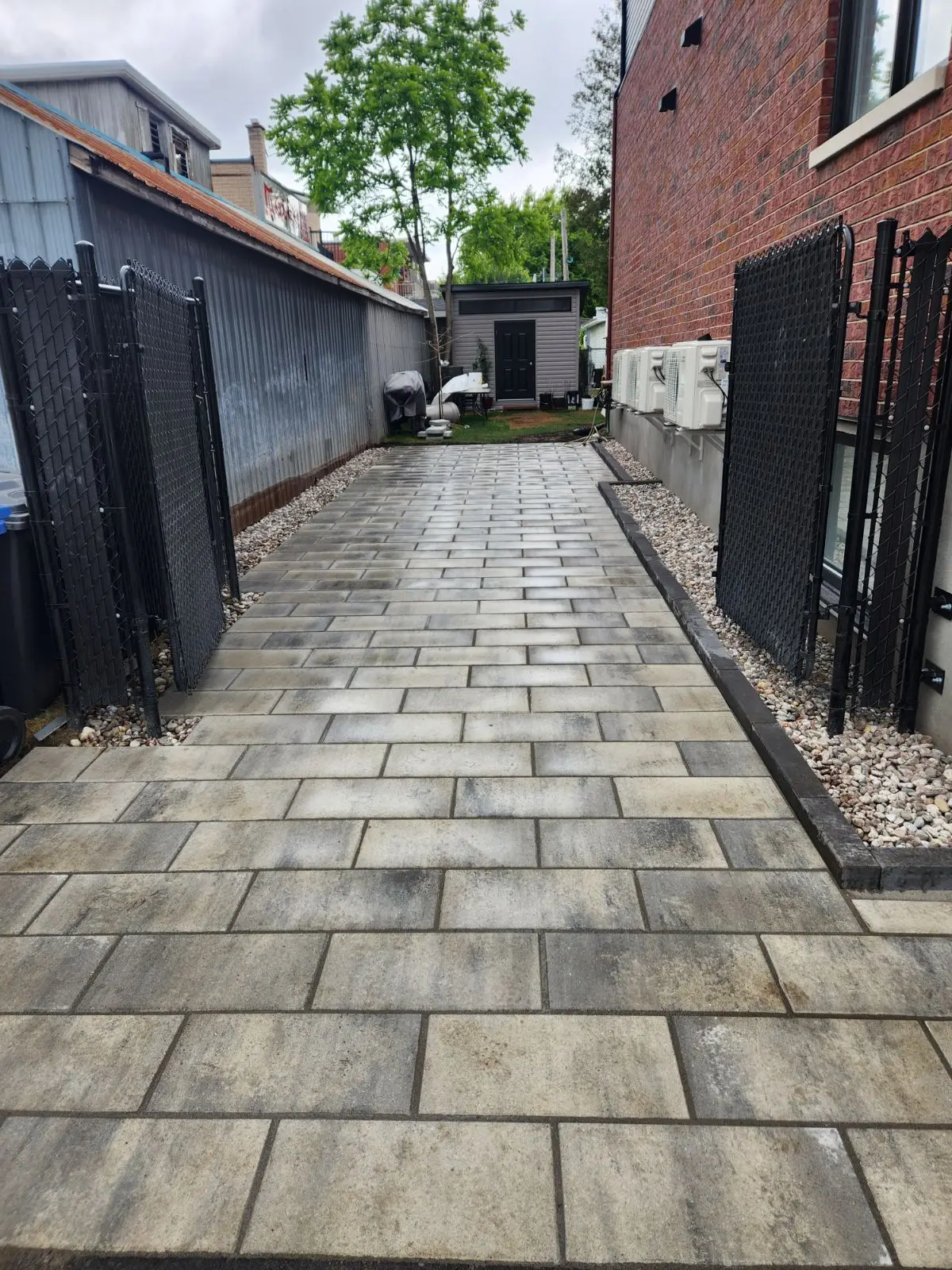 Outdoor walkway with grey interlocking pavers between two buildings, black edging, metal fences, and decorative stones along the sides.