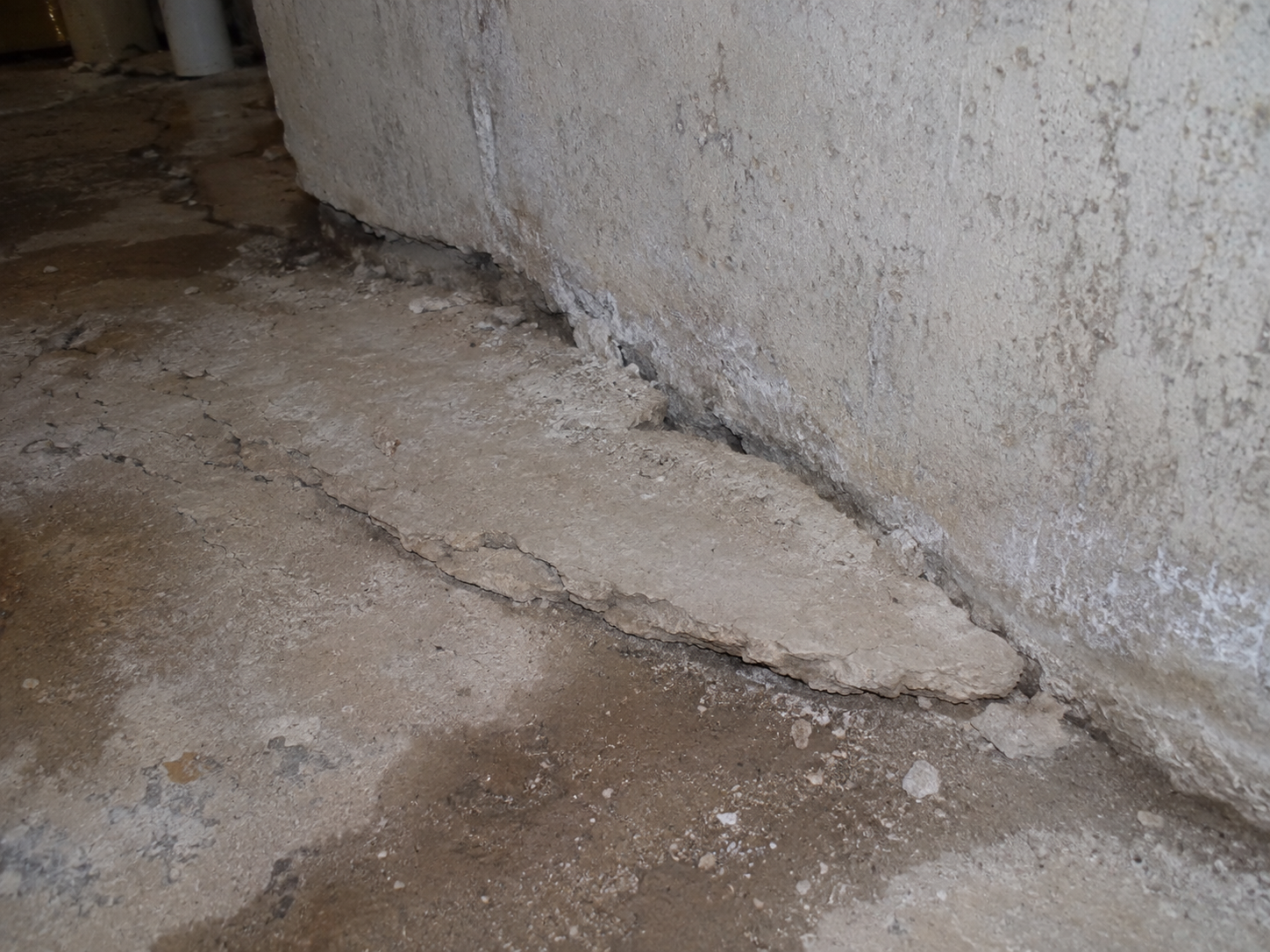 Une vue rapprochée d’un plancher de sous-sol où le béton s’effrite, se soulève et se fissure à la jonction avec le mur de fondation.