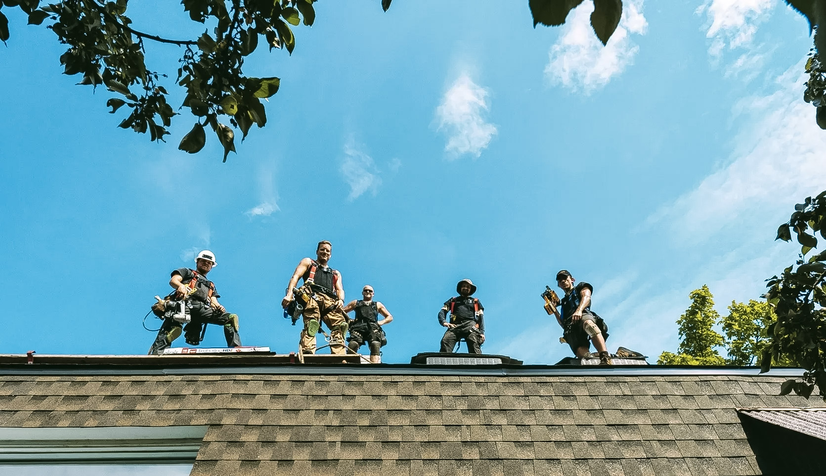 Cinq couvreurs munis de harnais de sécurité et d’outils sont debout sur le toit d’une maison résidentielle, sous un ciel bleu éclatant, entourés de branches d’arbres.