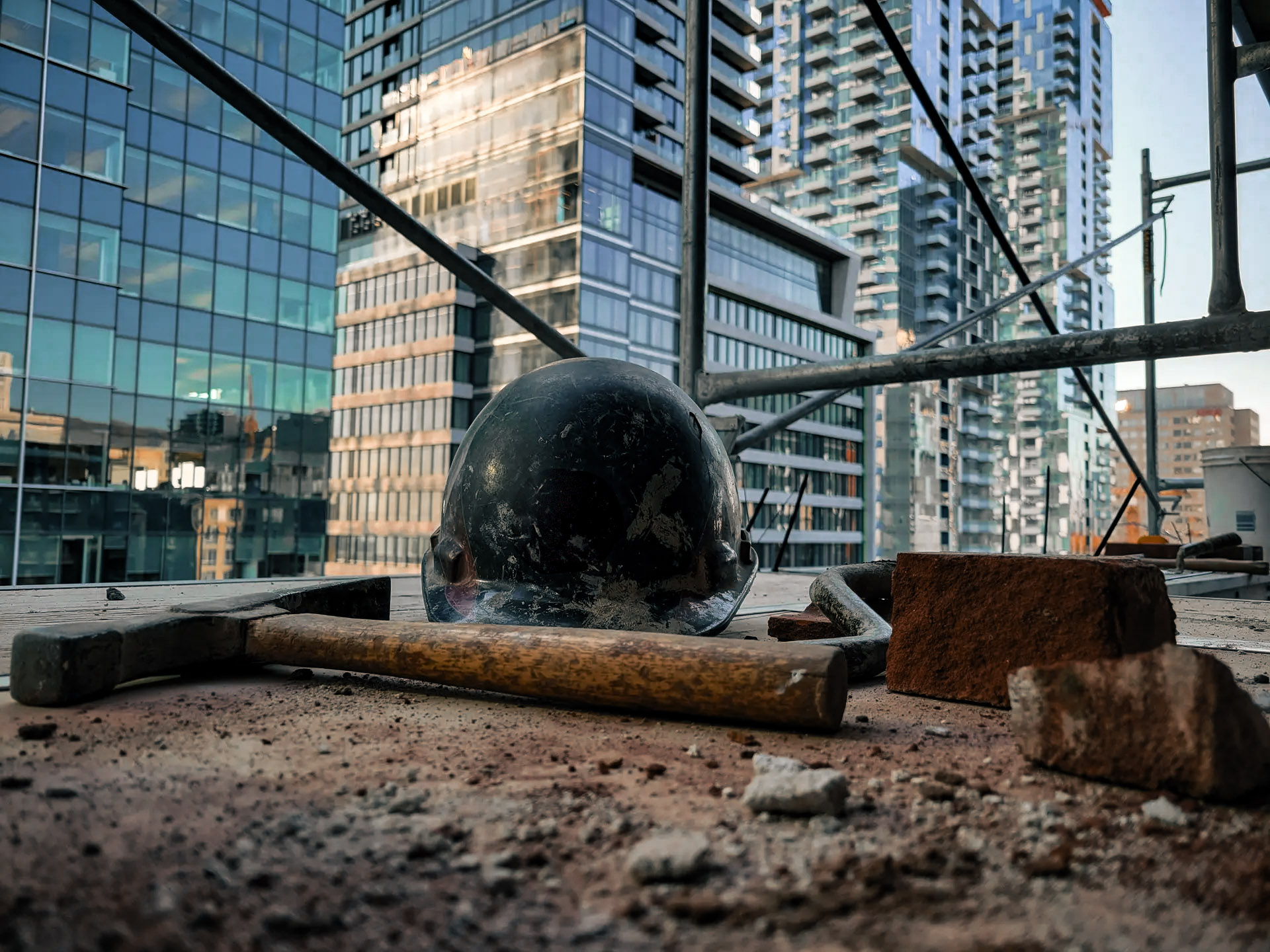 View of a high-rise construction site with a safety helmet, hammer, and bricks, surrounded by modern buildings in the background.