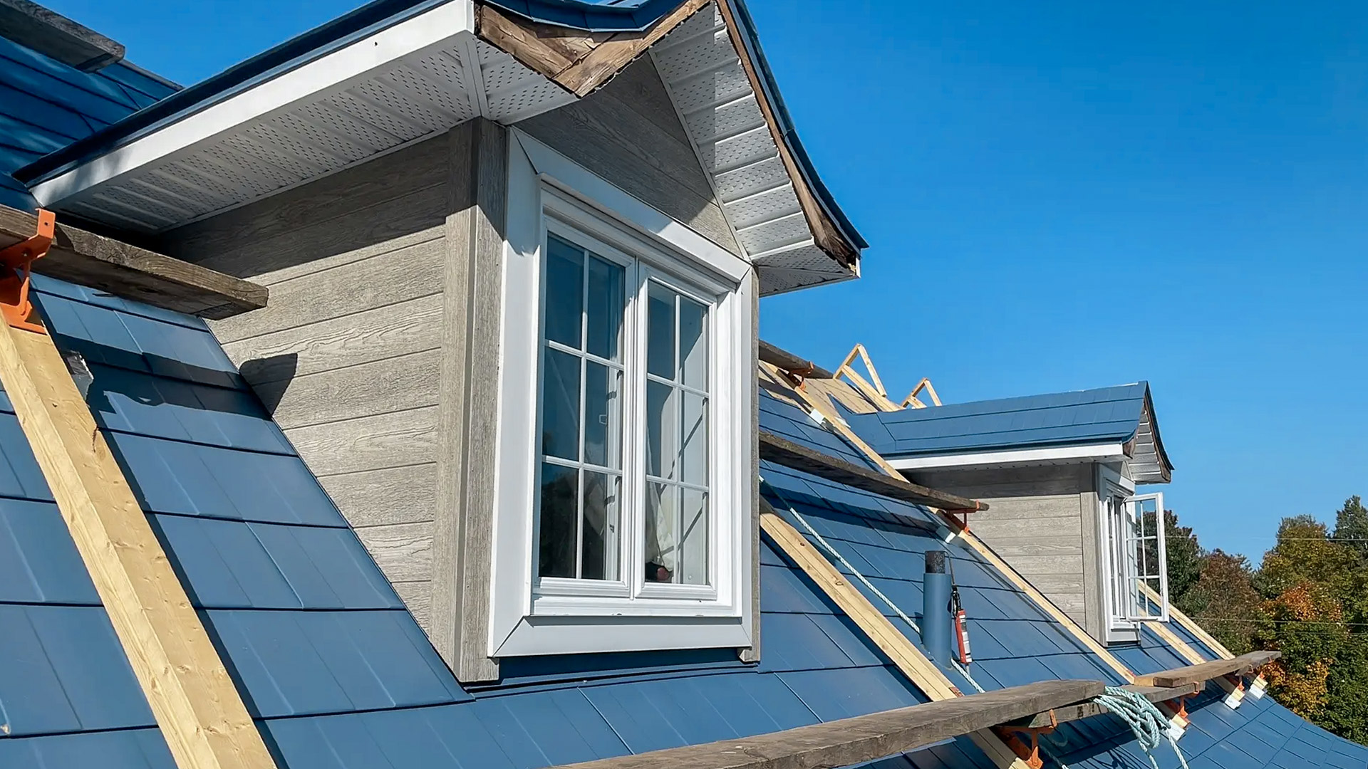 Installation of blue metal shingles on a roof with dormer windows and composite wood siding