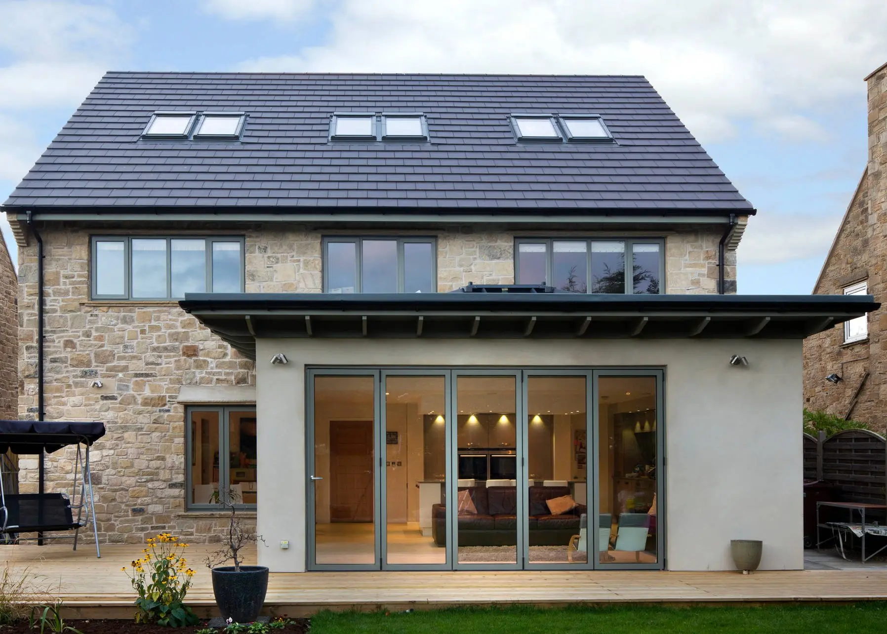 Maison en pierre rénovée avec toiture en tuiles, fenêtres à double vitrage, portes-fenêtres pliantes, terrasse en bois et éclairage extérieur moderne.