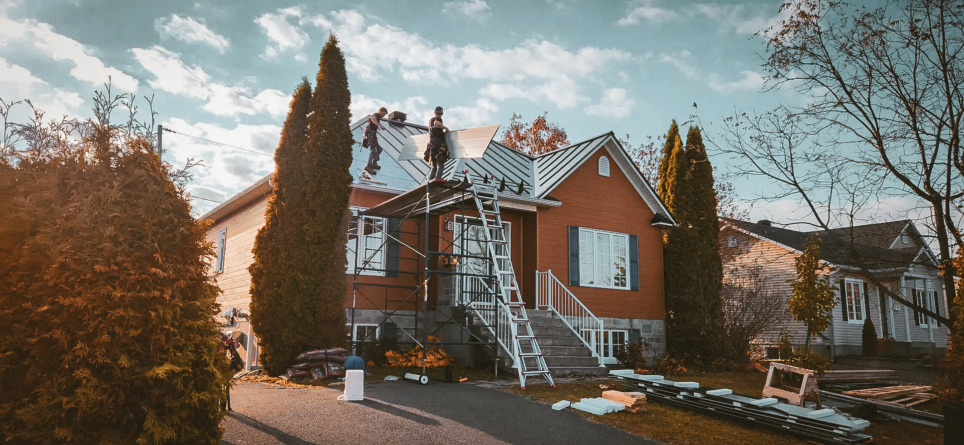 Roof renovation work on a residential house with workers installing metal panels, scaffolding, and equipment on site.
