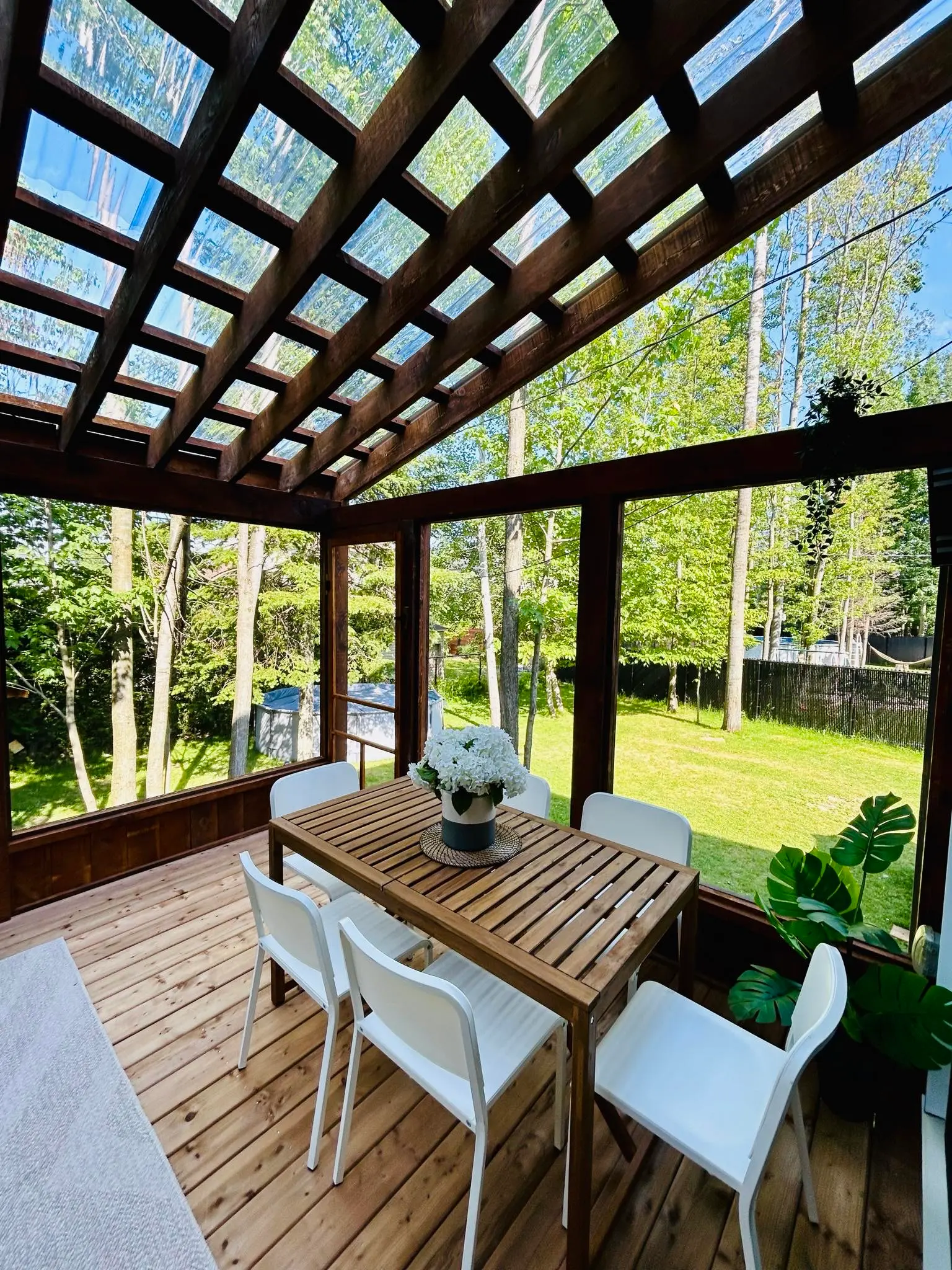 Véranda en bois avec toit en verre, table de jardin en bois et chaises blanches, offrant un espace extérieur lumineux et moderne entouré de verdure.