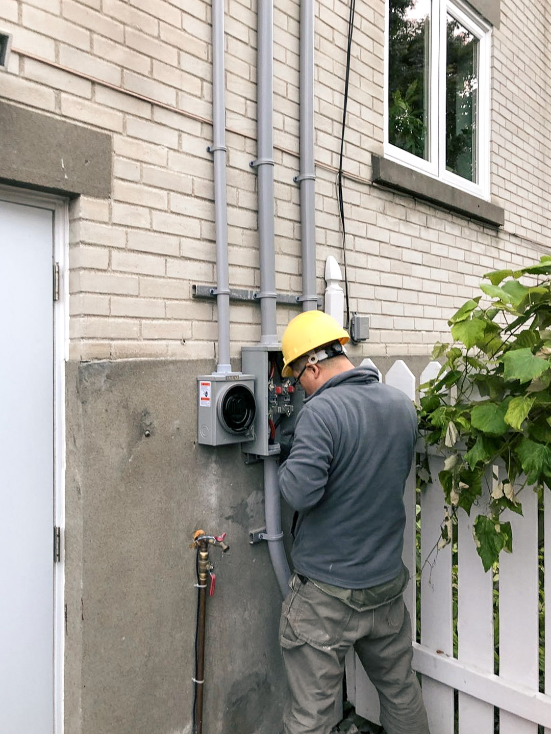 Electrician in yellow helmet performing outdoor electrical installation on beige brick wall with gray box and conduits
