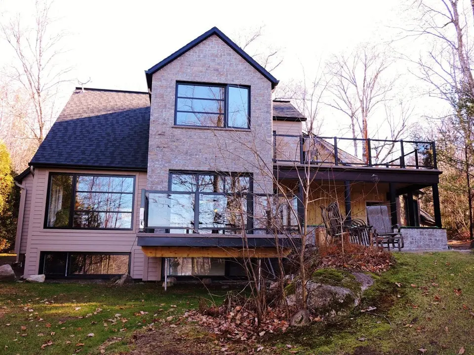açade arrière d’une maison contemporaine en brique et revêtement beige avec grandes fenêtres, terrasse surélevée, garde-corps en verre et balcon supérieur