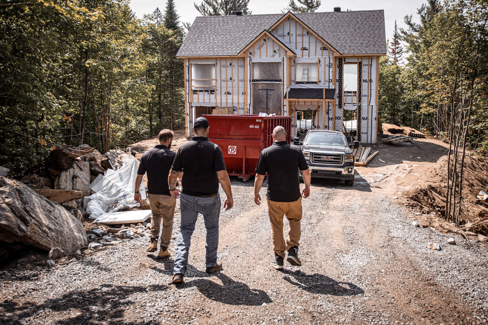 Three men in black shirts and beige or blue pants walk toward a house under construction, surrounded by trees, with a red dumpster and a pickup truck in front of the home.
