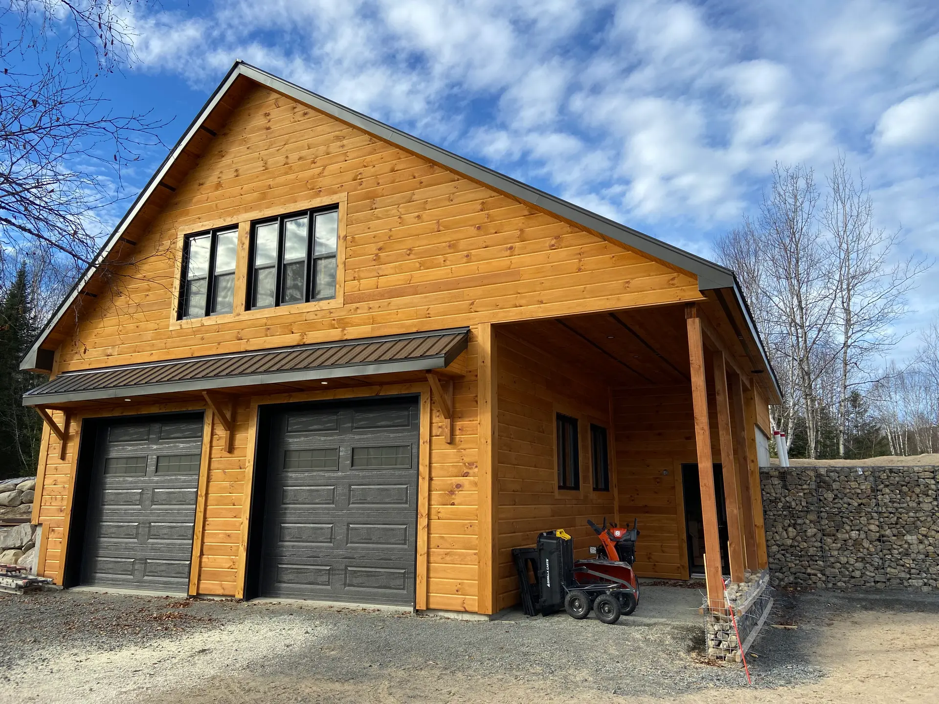 Garage double en bois avec revêtement naturel, portes noires et abri latéral sur terrain résidentiel avec mur de pierres