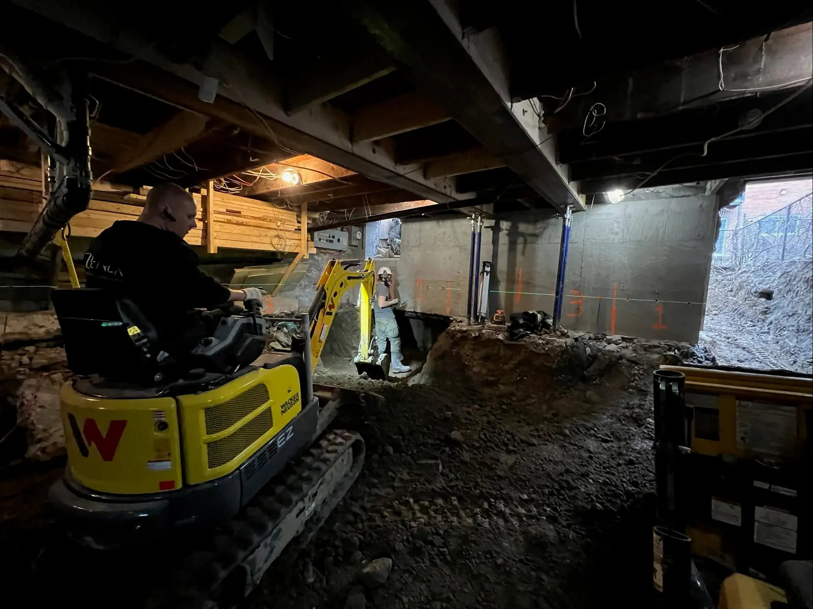 Excavation work beneath a house with mini excavator, workers, concrete foundation, exposed soil, and visible floor structure