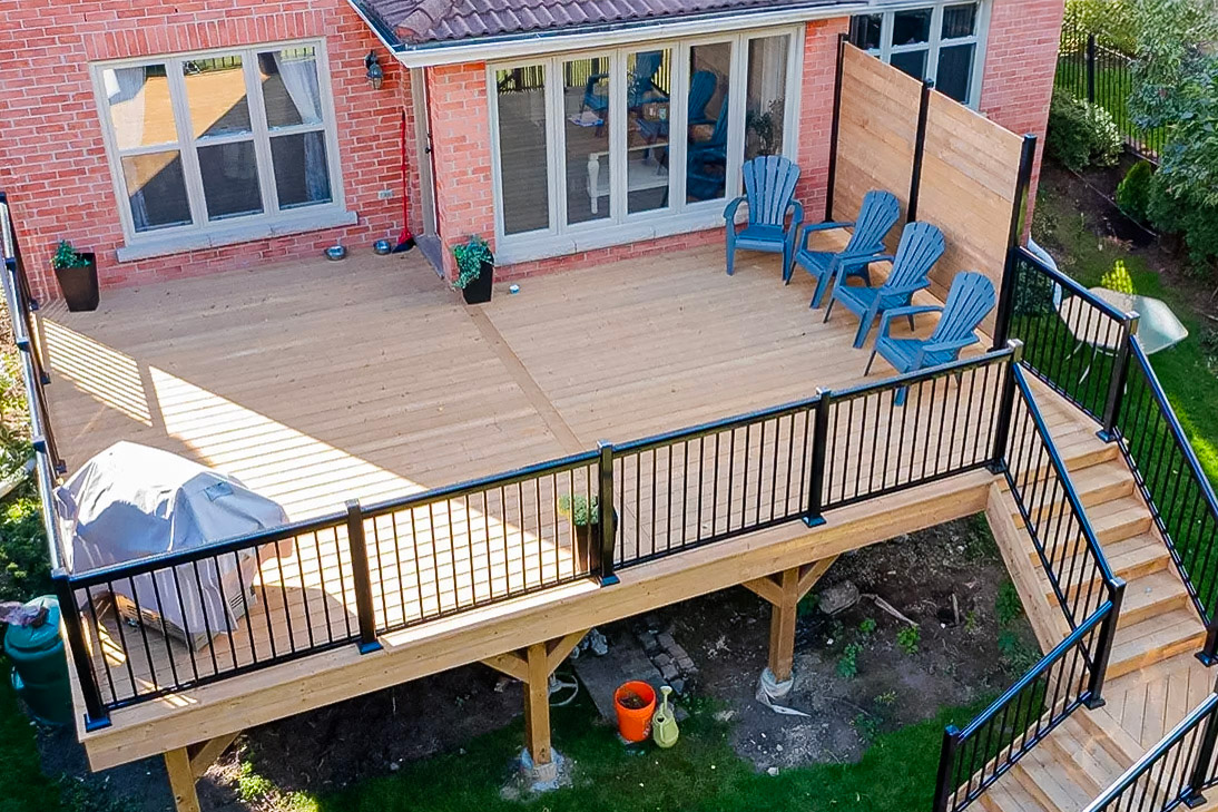 Elevated wooden deck with black metal railing, garden chairs, covered barbecue, and staircase leading to the garden, attached to a red brick house.