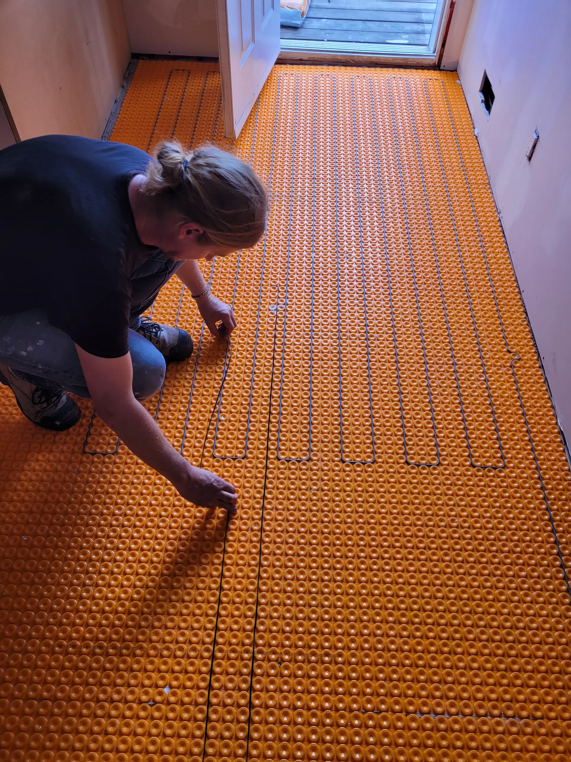 Worker installing an electric underfloor heating system with an orange membrane in a room under construction.