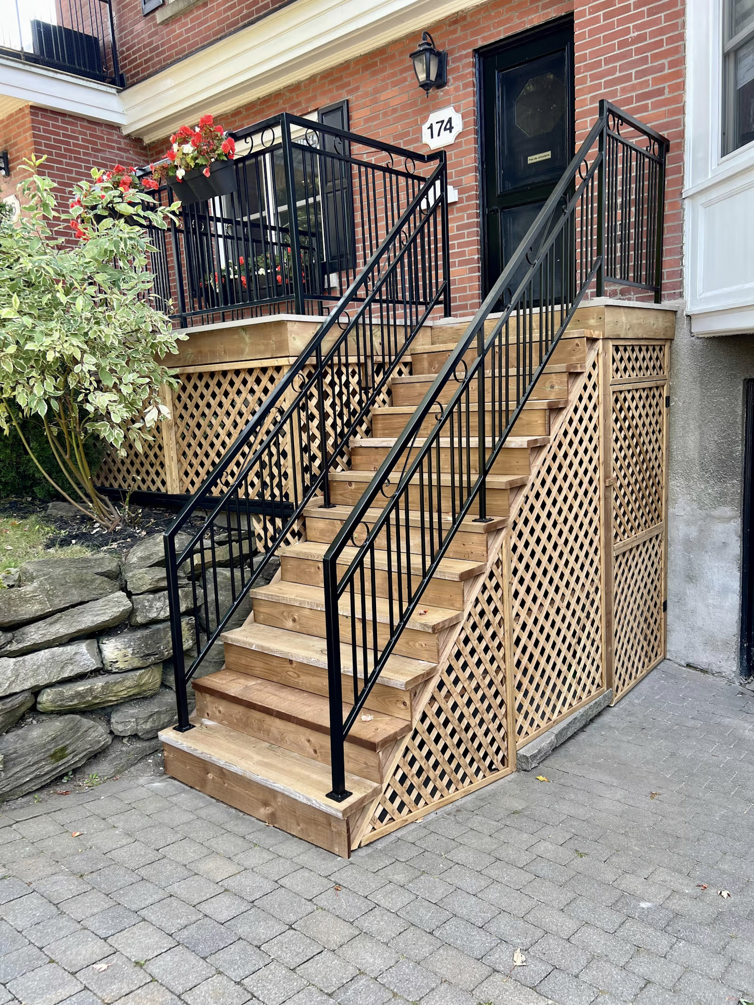 Outdoor wooden staircase with black metal railings, a raised landing, decorative lattice, and integrated storage in front of a brick house.