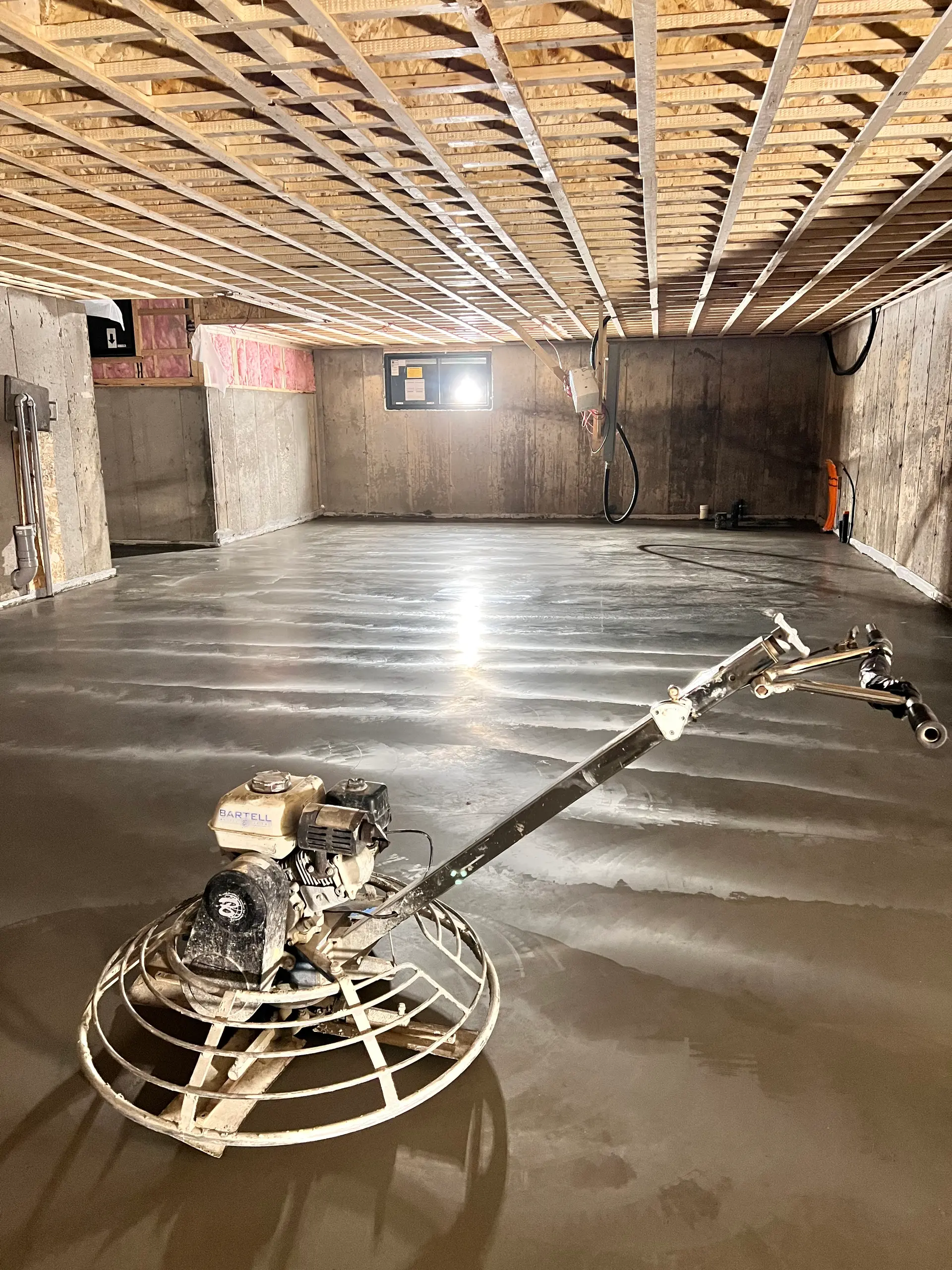 Basement renovation with freshly smoothed concrete slab, power trowel, exposed foundation walls, and visible floor joists