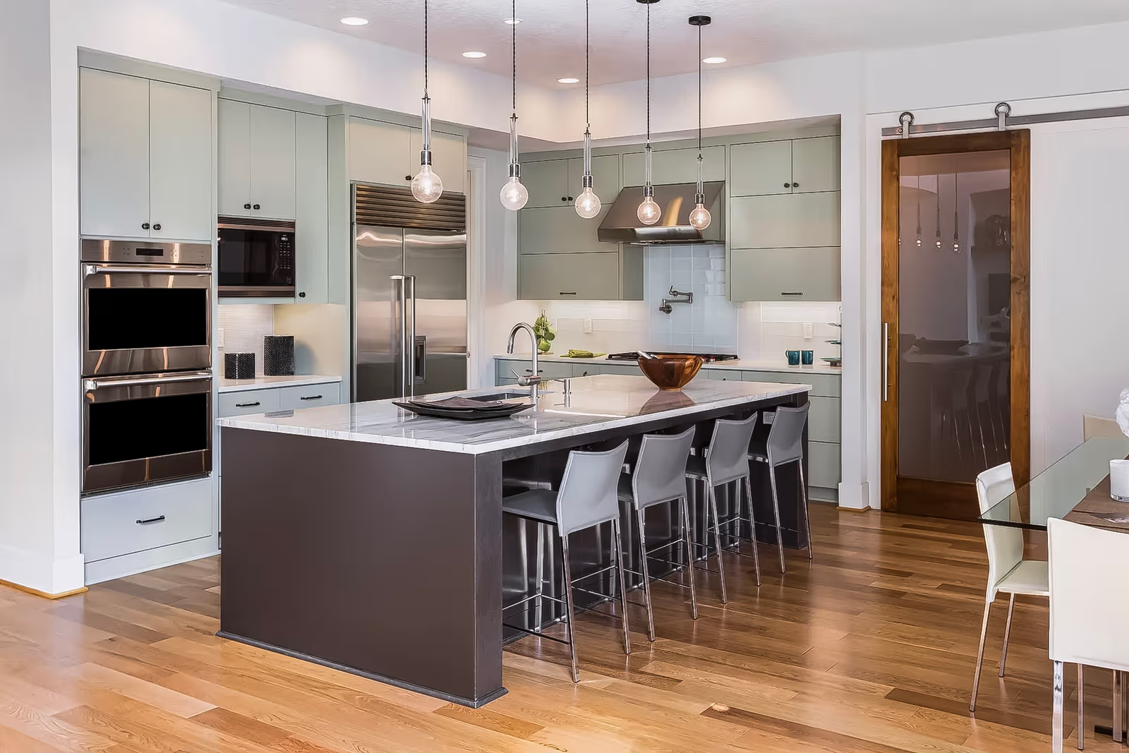 Cuisine moderne avec îlot central en quartz, armoires vert sauge, électroménagers encastrés en acier inoxydable, luminaires suspendus et plancher de bois franc.