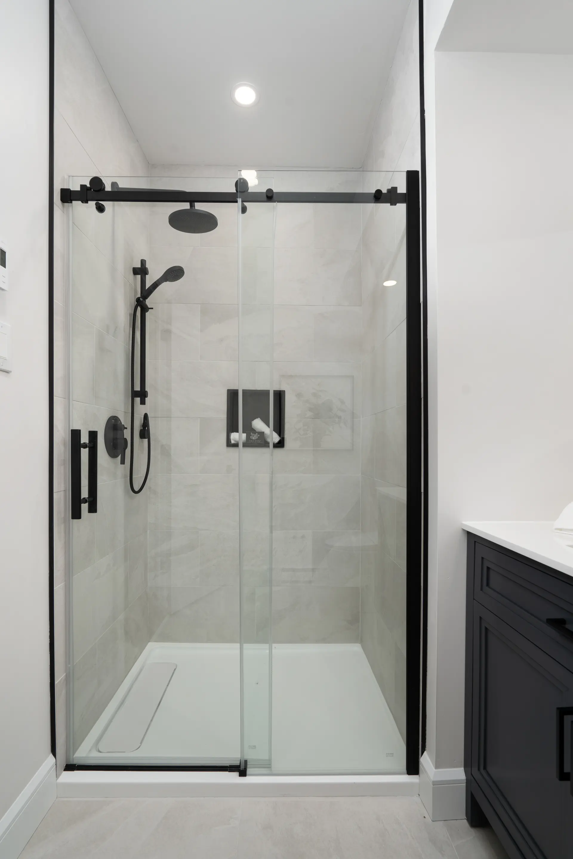 Modern shower with a sliding glass door, black hardware, white base, light marble-effect tiles, and a black vanity.
