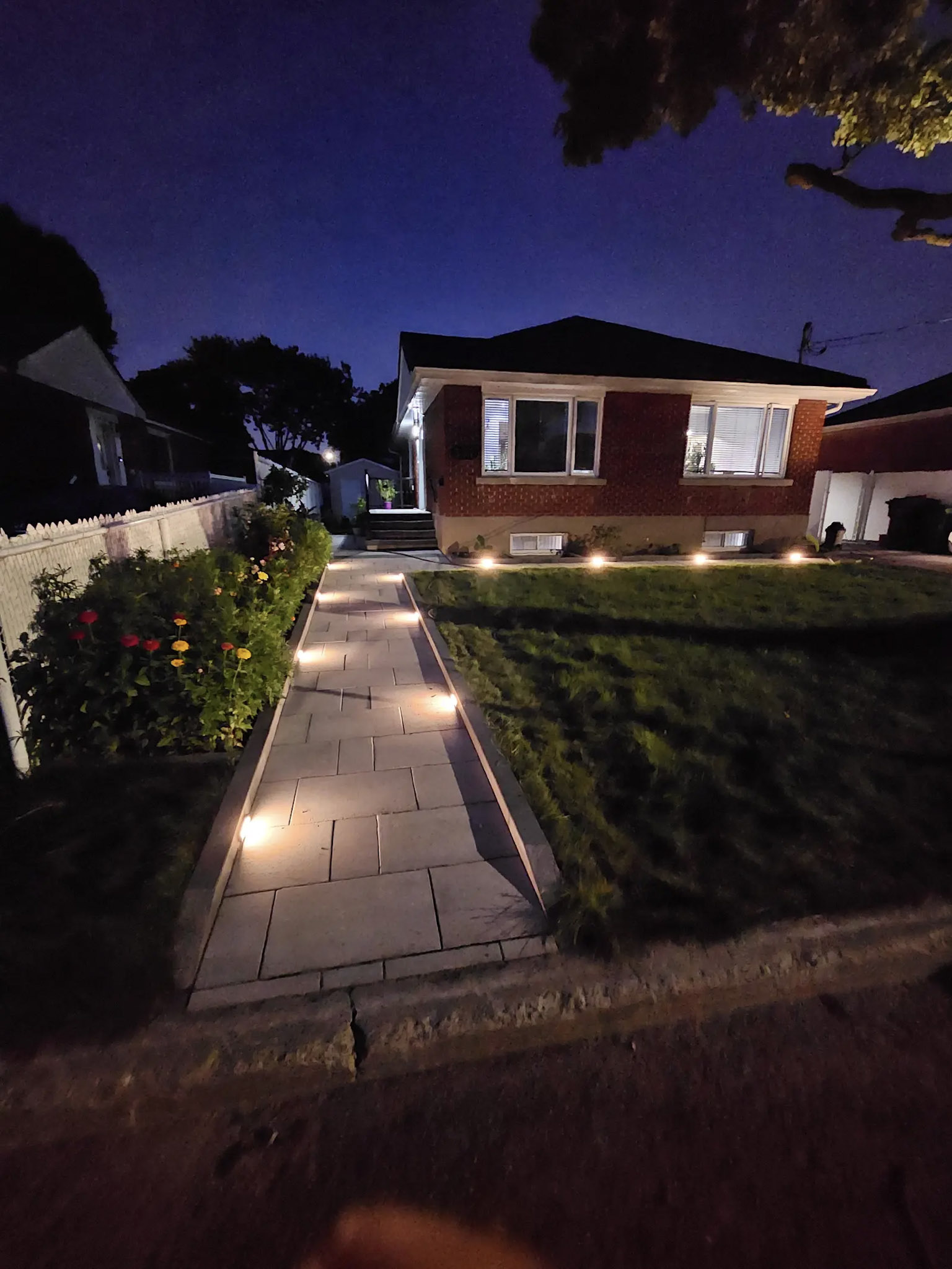 Brick house illuminated at night with paver walkway, landscape lighting and flower garden.