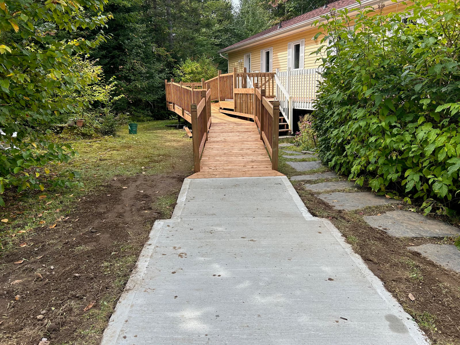Wooden accessibility ramp with railings leading to a yellow house surrounded by greenery with a concrete walkway entrance