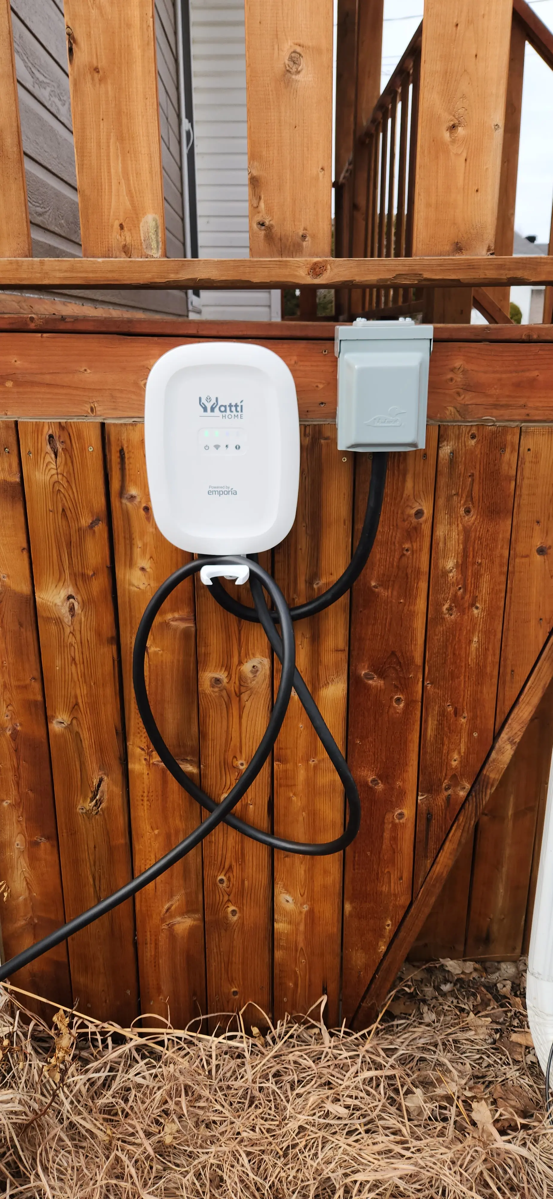 Wall-mounted electric vehicle charging station installed on outdoor wooden fence with electrical outlet and visible cable