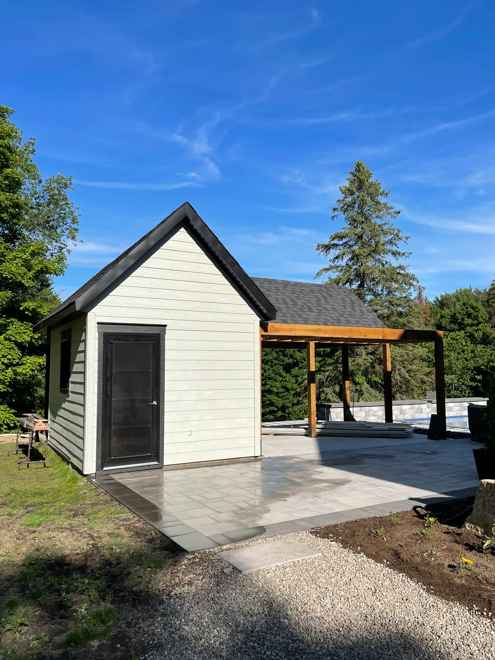 Modern shed with white siding, black door, shingle roof, slab patio, wood pergola, low wall and outdoor landscaping in progress