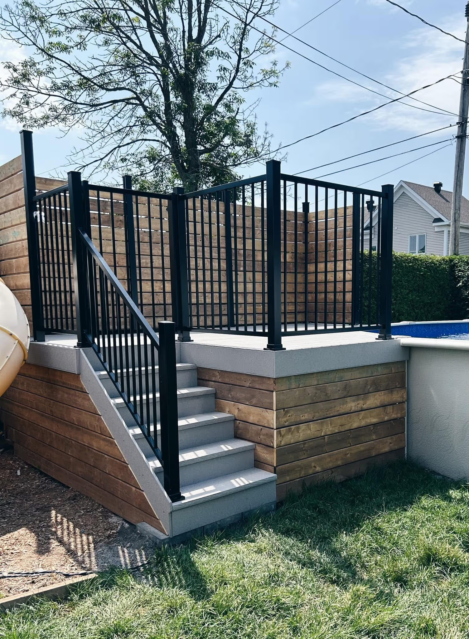 Terrasse surélevée avec escalier gris, garde-corps noir en aluminium et panneaux d’intimité en bois près d’une piscine résidentielle