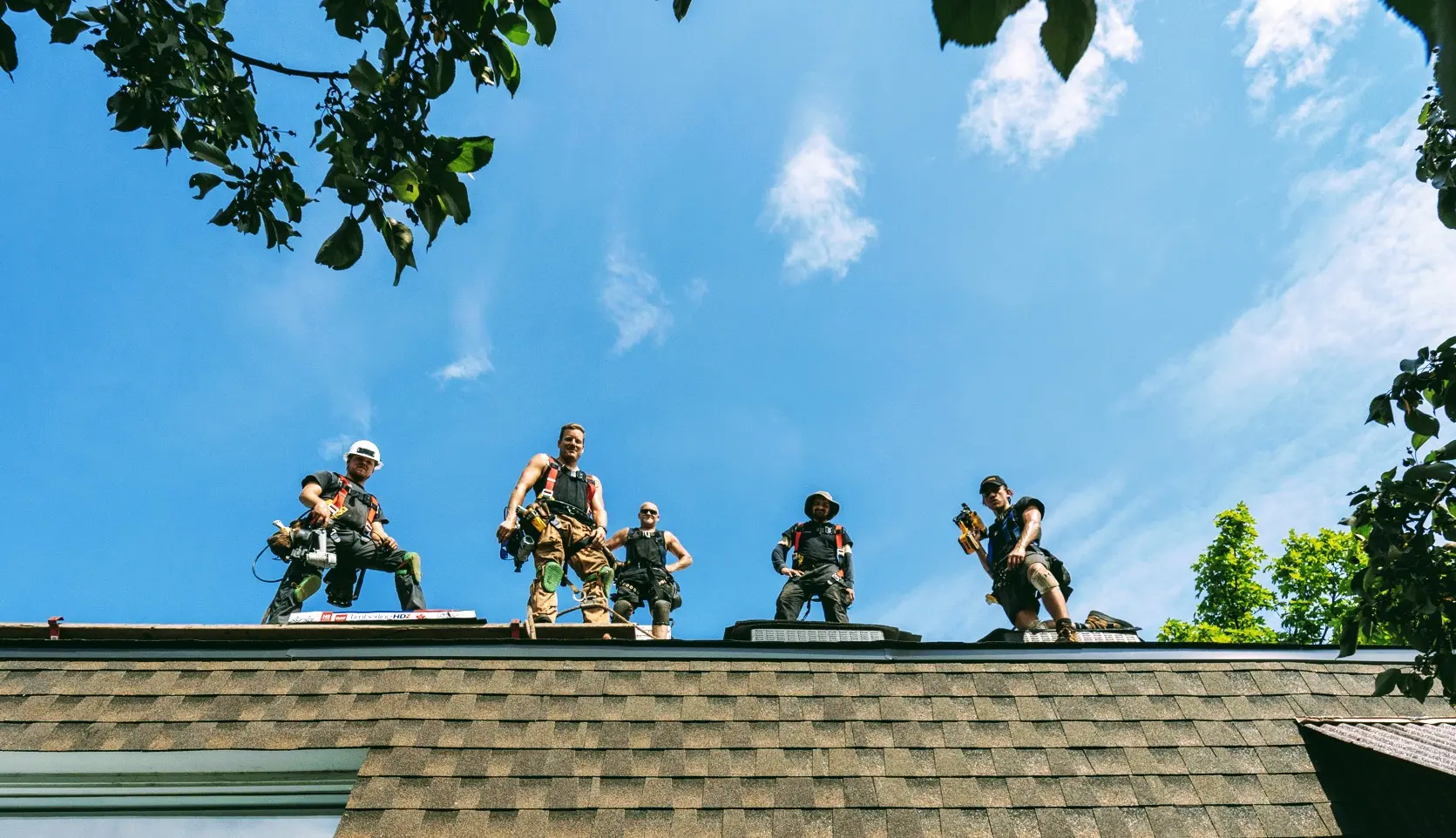 Team of roofers working on a roof under a blue sky, wearing harnesses and construction tools.
