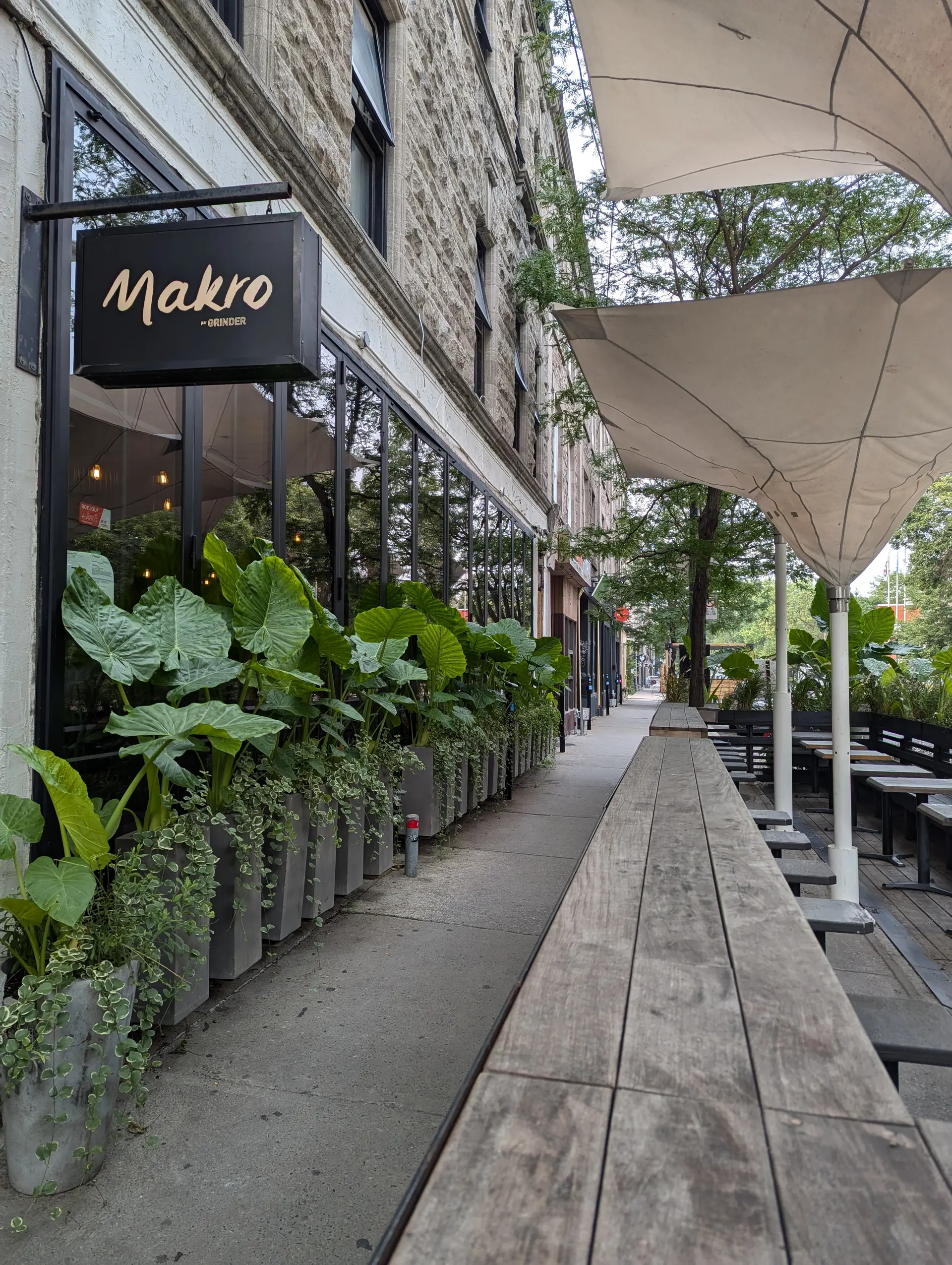 Terrasse extérieure de restaurant avec longues tables en bois, parasols, plantes en bacs et façade en pierre sur une rue urbaine.