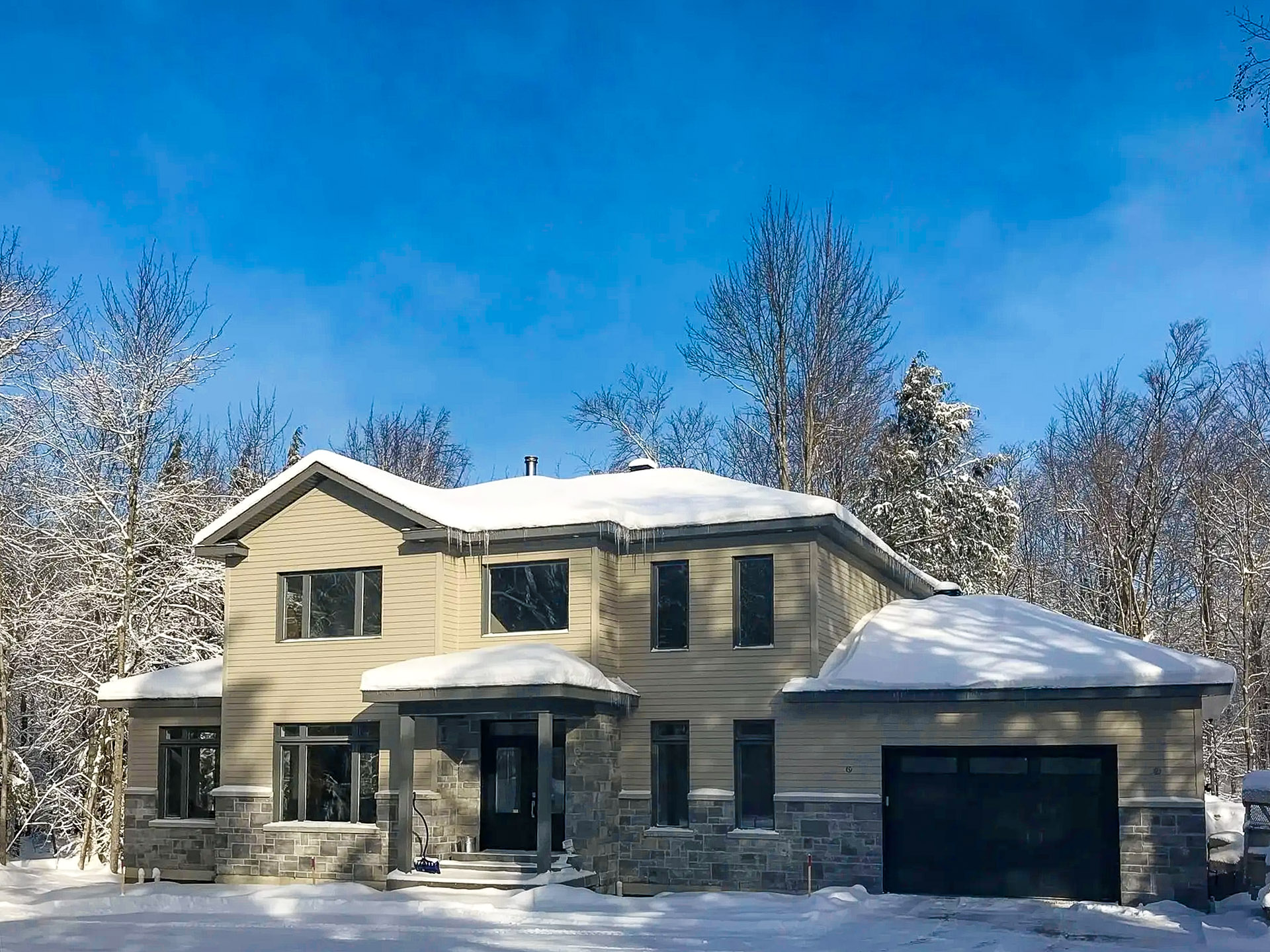 Contemporary house with beige stone and vinyl siding, surrounded by snow-covered trees and a snow-covered roof