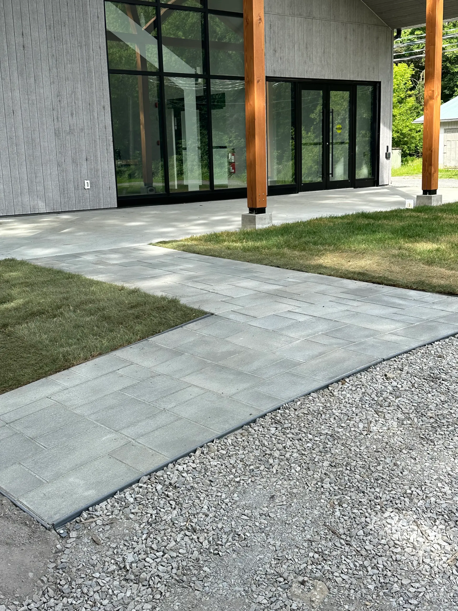 Allée en pavé uni gris menant à une maison moderne avec grandes portes vitrées, revêtement gris, colonnes en bois et gravier décoratif.