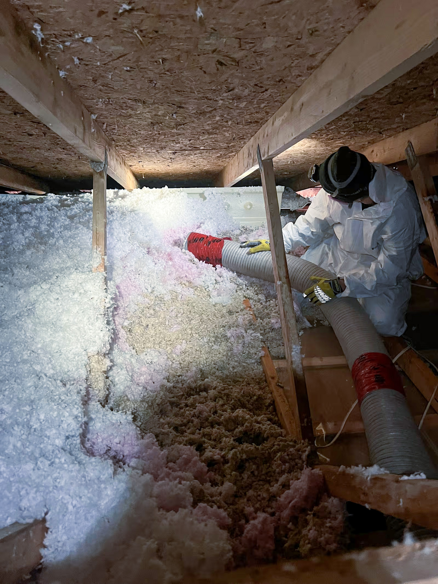Technicien en combinaison de protection installant de l’isolant soufflé dans un entretoit résidentiel avec boyau flexible et charpente de bois.