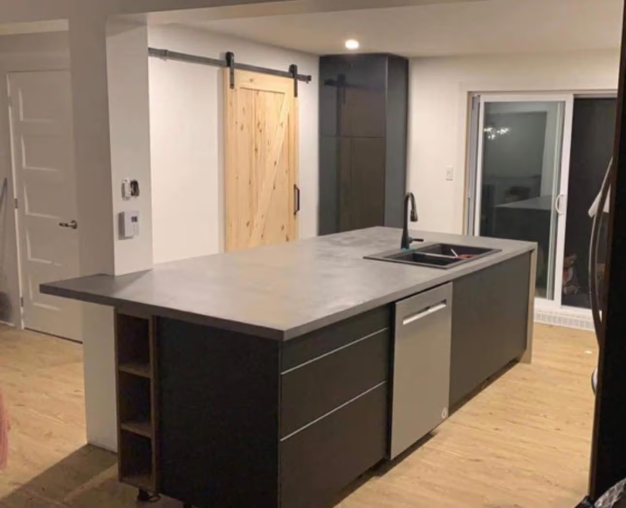 Cuisine moderne avec grand îlot gris, armoires noires, évier intégré, robinet noir, porte coulissante en bois et plancher clair