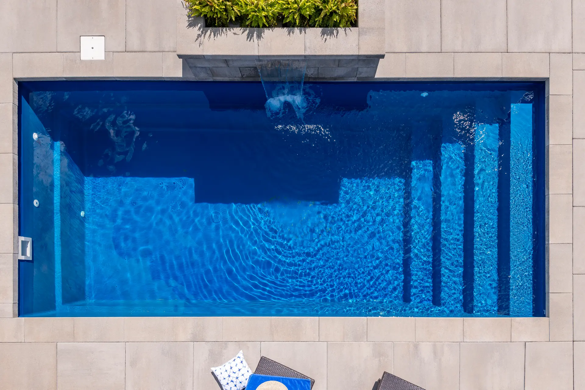 Vue aérienne d'une piscine rectangulaire avec cascade intégrée, entourée de dalles modernes et de végétation décorative.&nbsp;