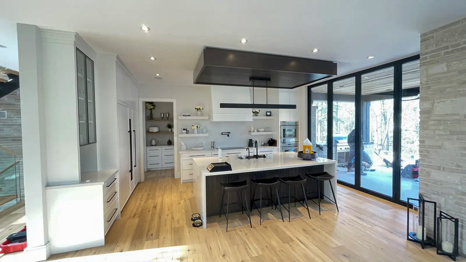 Cuisine contemporaine lumineuse avec grand îlot central, comptoir blanc, tabourets noirs, armoires épurées, plancher en bois et larges portes vitrées donnant sur la terrasse.