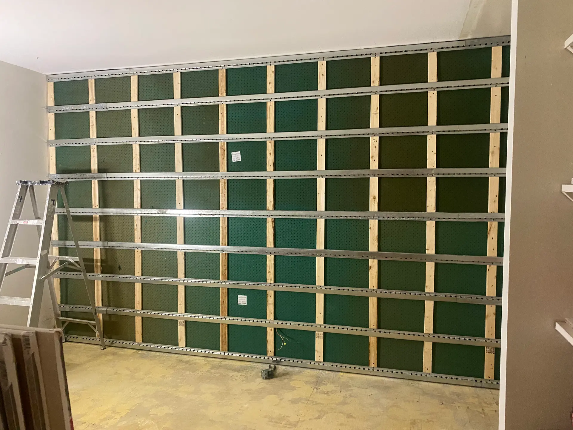 Interior wall under renovation with wood framing, horizontal metal rails, green panels and construction ladder
