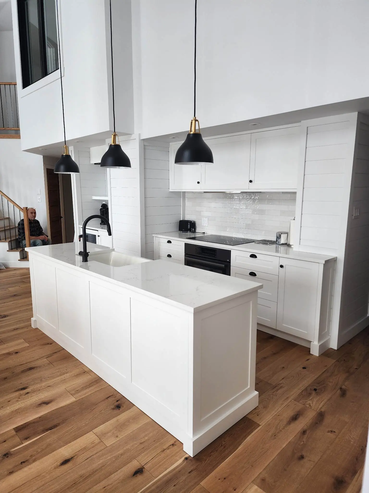 Modern white high-ceiling kitchen with large central island, white countertop, shaker cabinets, black pendant lights, black faucet, subway tile backsplash and wood flooring