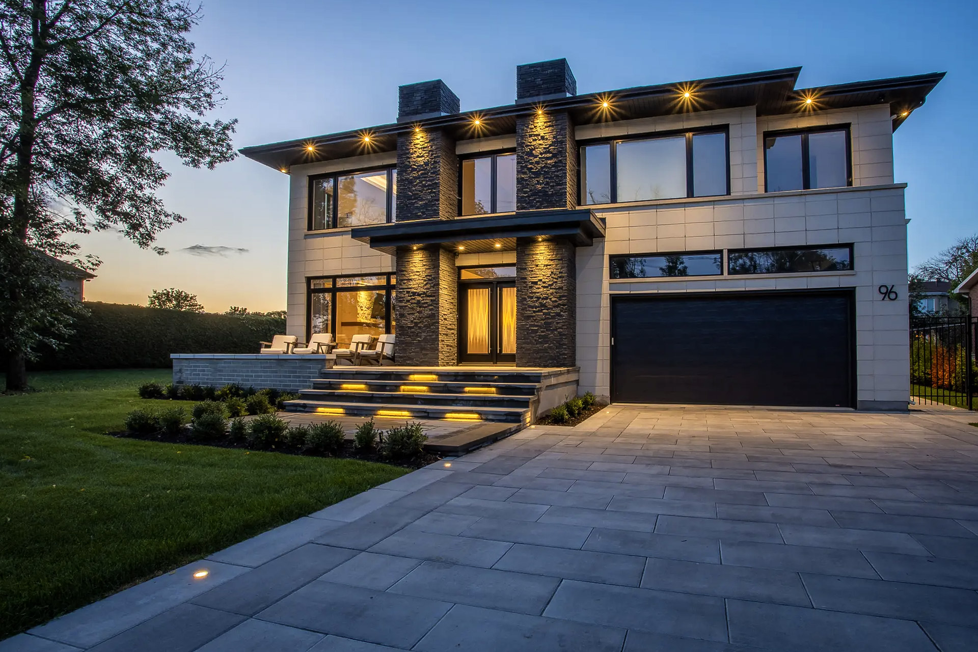 Modern contemporary style house with integrated exterior lighting, stone facade and light panels, black garage door, and large gray paving stone entrance, surrounded by greenery.