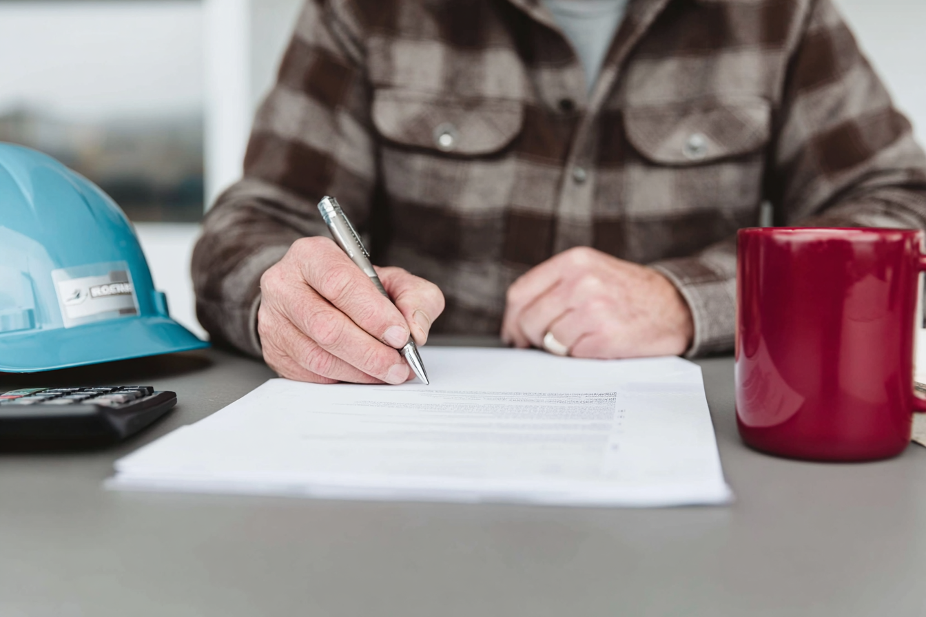 Personne en chemise à carreaux signant un contrat ou document de chantier avec un casque de sécurité bleu et une tasse rouge sur la table.