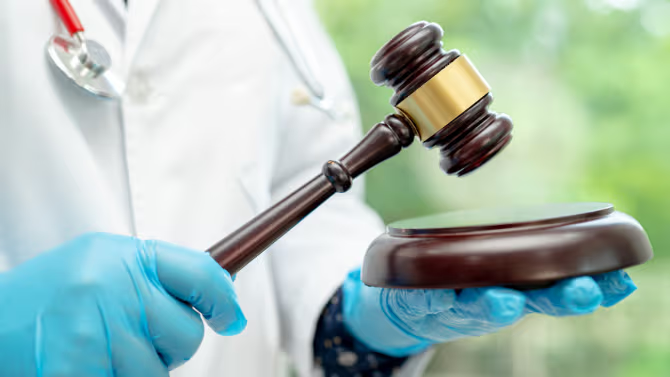 A person wearing a medical coat and blue gloves holds a judge’s gavel above a sound block, symbolizing legal, regulatory, or litigation matters in the healthcare field.