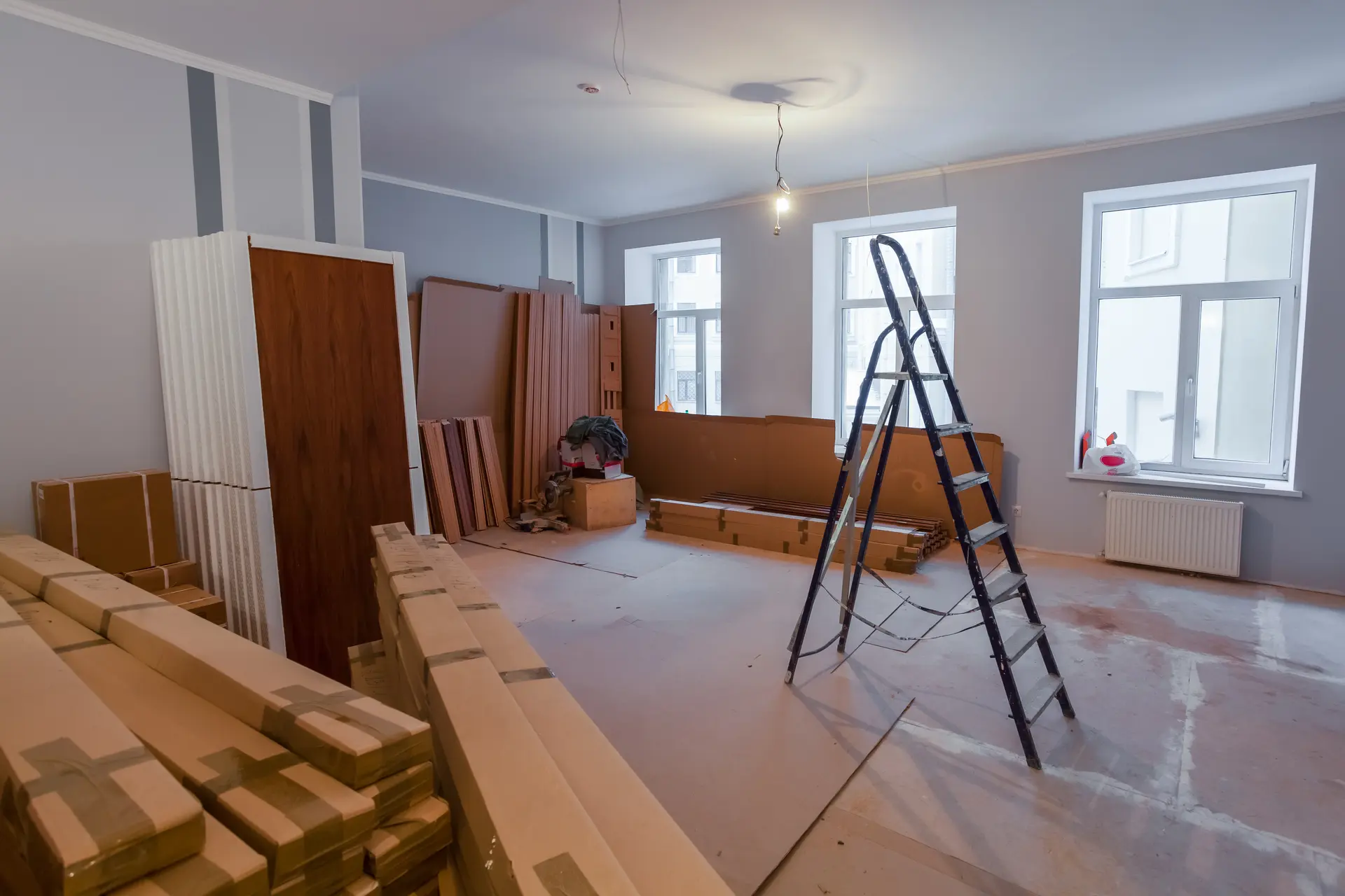 Interior room under renovation with freshly painted gray walls, a ladder, stacked construction materials, white windows, and exposed ceiling wiring.