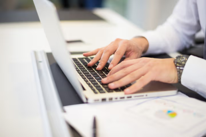 Hands of a professional typing on a laptop with work documents on a modern office desk.