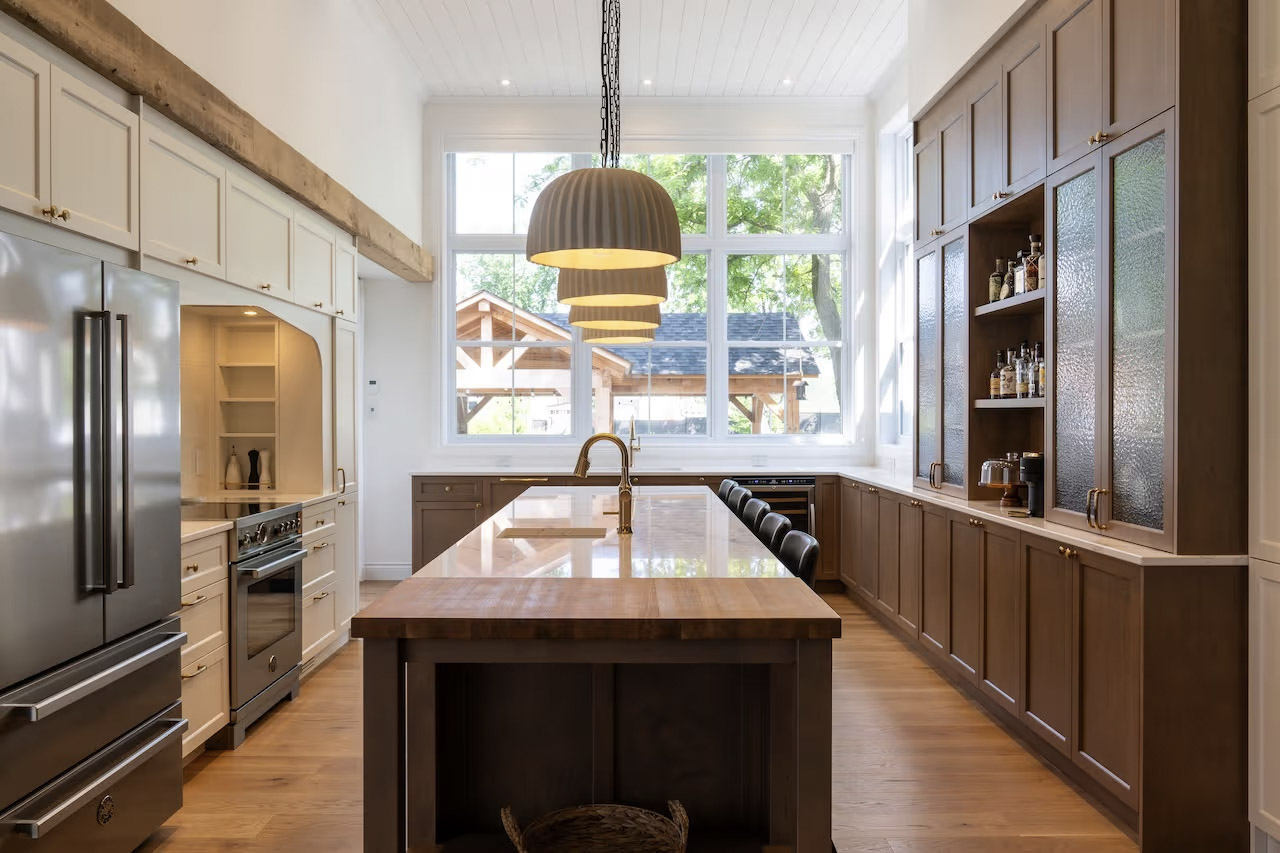 Bright kitchen with a large central island, white and brown cabinetry, stainless steel appliances, and tall windows.