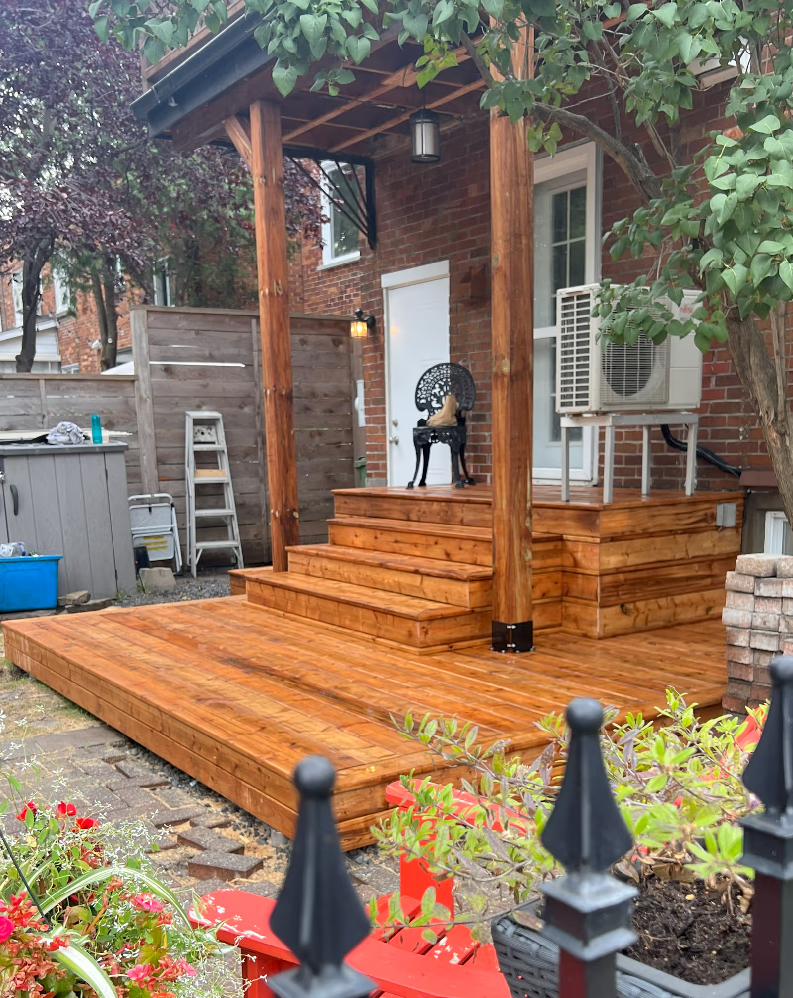 Wood deck with stairs, platform, covered roof, and posts beside a brick house, surrounded by trees, plants, and a fence