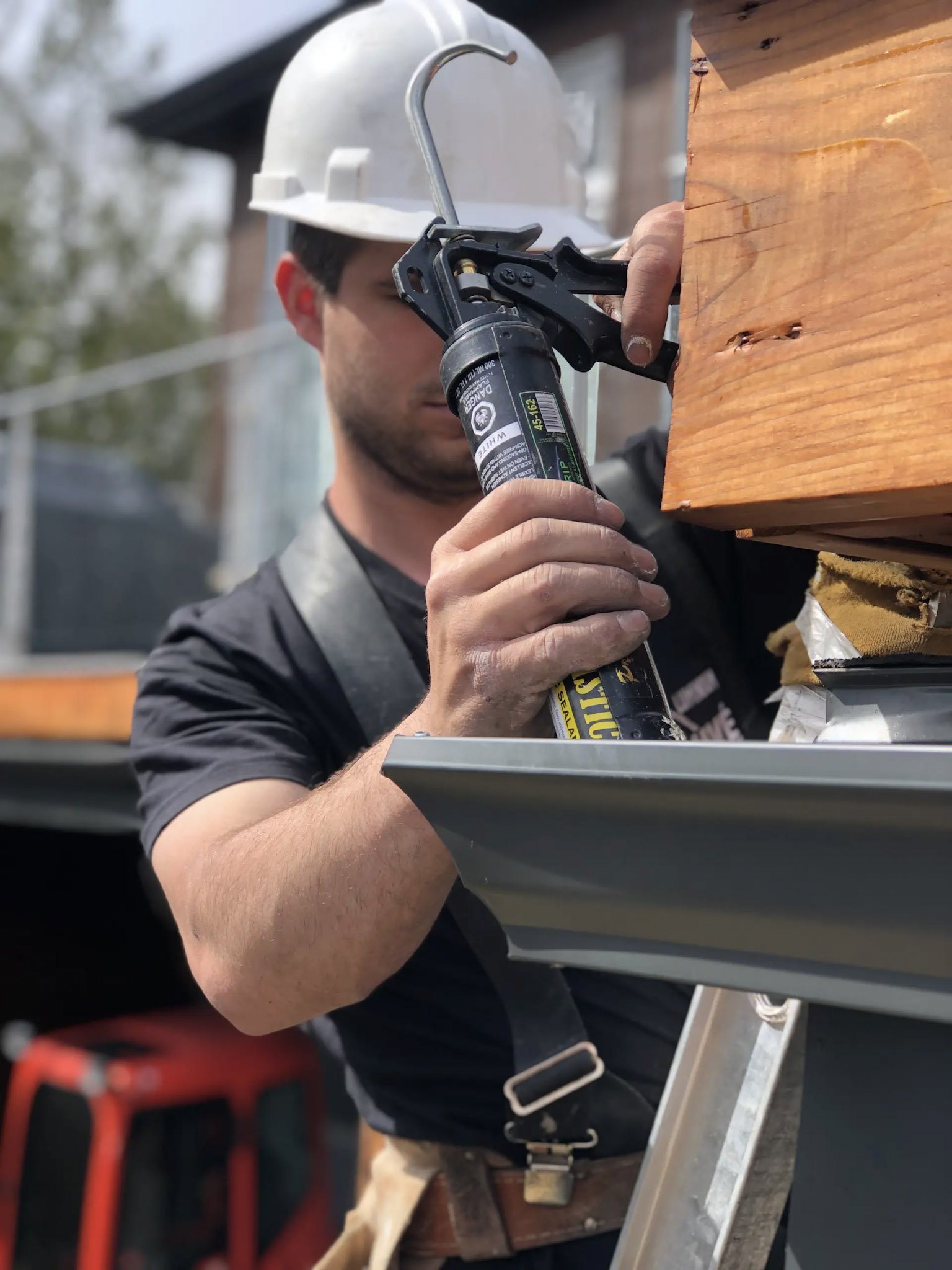 Travailleur de la construction portant un casque blanc et appliquant du scellant avec un pistolet à calfeutrer sur une structure en bois près d’une gouttière extérieure.